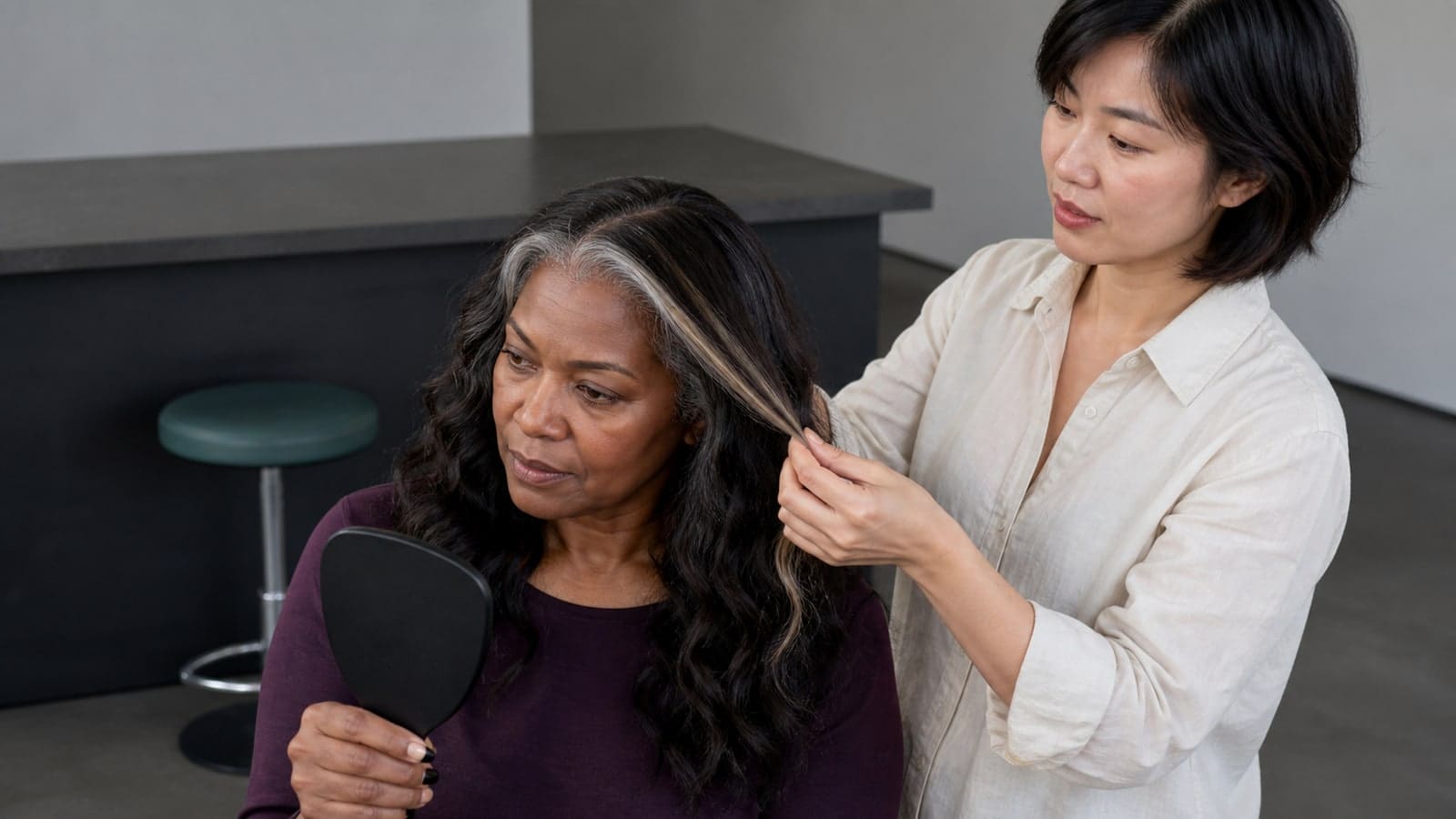 Colorist separates a narrow icy face-framing ribbon from a client's glossy dark brunette waves while the client checks the placement in a handheld mirror