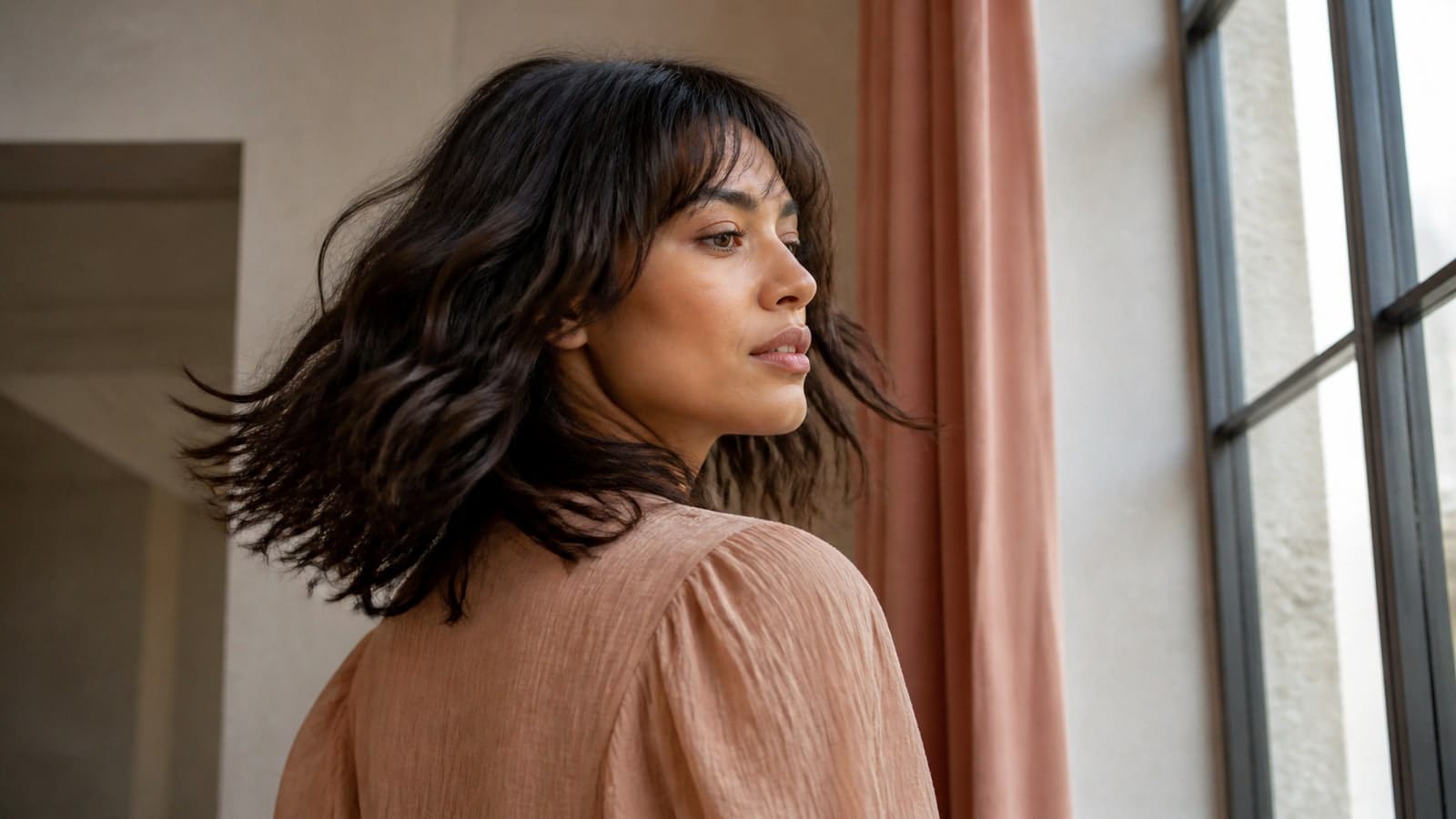 A woman turning beside a window with a dark espresso shoulder-length wavy shag, curtain fringe, cheekbone face framing, short crown layers, and a softly broken perimeter